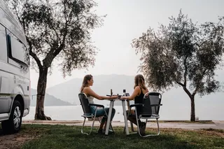 Two women sit on a camping set in front of a VW Grand California.