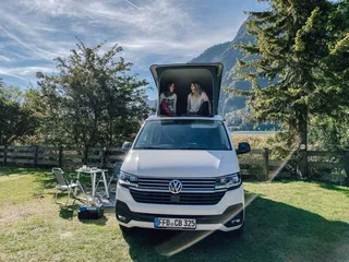 Two women sit in the open roof of a VW bus in nature.