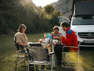 Eine Freundesgruppe sitzt um einen Campingtisch beim Frühstück beim Camping im Münstertal. Im Hintergrund ein Mercedes Marco Polo Camper.