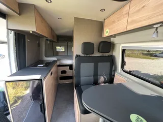 Geräumiger Innenraum eines Campervans mit Blick auf die Küchenzeile, die gemütliche Sitzbank und das feste Bett hinten.