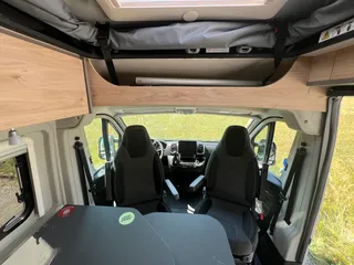 Blick auf das Cockpit eines modernen Campers mit umgedrehten Pilotensitzen und dem großzügigen Bettbereich im geöffneten Aufstelldach.