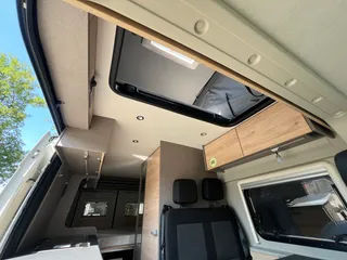 Innenraum eines modernen Campervans mit Blick auf die gemütliche Sitzbank, Holzschränke und die Deckenöffnung zum Schlafdach.