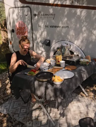 Eine Frau sitzt an einem Campingtisch mit gedecktem Frühstück vor der Tür eines weißen Vanever Wohnmobils Modell Corsica.