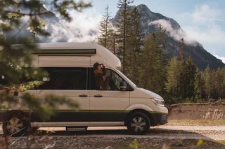 A camper is in a forest. A woman looks out of the window.