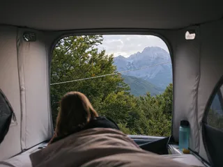 A woman is in a camper in bed and looks at a forested mountain landscape.